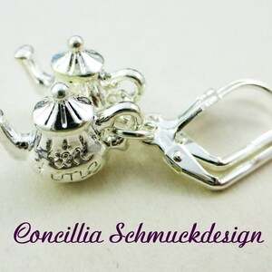 Könnte beinhalten: Silberfarbene Teekannen-Ohrringe mit filigranen Details. Die Ohrringe haben einen Klappverschluss und sind wie Miniatur-Teekannen mit Deckeln und Henkeln geformt. Der Text "Concillia Schmuckdesign" ist im Bild zu sehen.