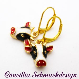 Könnte beinhalten: Ein Paar goldfarbene Ohrhänger mit Kuhkopf-Anhängern. Jeder Anhänger ist mit weißer, schwarzer und roter Emaille verziert und zeigt ein Kuhgesicht mit weißer Schnauze, schwarzen Flecken sowie roten Ohren und Nüstern. Darunter steht Concillia Schmuckdesign.