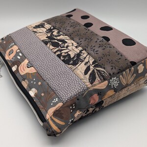 Könnte beinhalten: Eine graue und braune Patchwork-Tasche mit Reißverschluss und Blumendruck. Die Tasche hat einen schwarzen Reißverschluss und einen silbernen Reißverschluss-Zipper.
