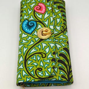 Könnte beinhalten: Eine grüne und schwarze gemusterte Geldbörse mit einem goldenen Verschluss und einem blauen und gelben Blumenanhänger. Die Geldbörse hat ein blaues Futter und einen Klappverschluss.
