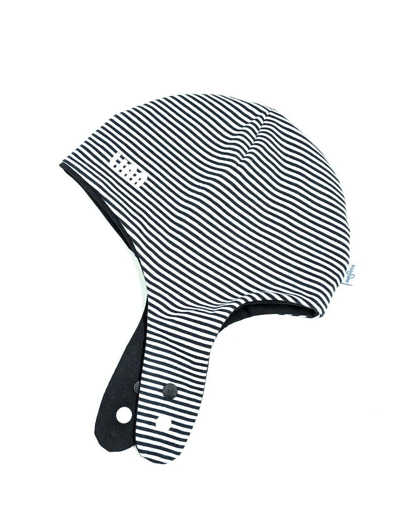 May include: A black and white striped hat with ear flaps, featuring a solid black interior lining and two black snap buttons on the flaps. The exterior fabric displays a repeating thin horizontal stripe pattern and includes the white text HAA.