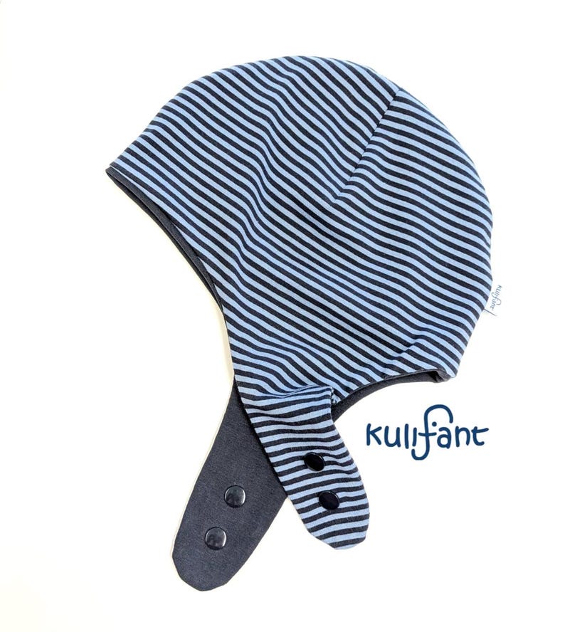 May include: A light blue and dark blue striped hat with ear flaps, featuring black snap buttons on the flaps and the text kulifant printed in dark blue. The hat has a rounded crown and a small white brand tag on the side seam.