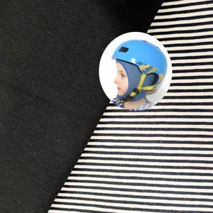 May include: A fabric swatch featuring a solid black section alongside a section with thin horizontal black and white stripes. A circular inset shows a blue helmet with yellow straps worn over a blue fabric head covering by a person in profile.