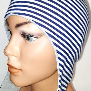 May include: A fitted cap featuring a pattern of thin horizontal navy blue and white stripes. The headwear has extended side flaps that frame the face, finished with a light grey trim along the edges and two visible circular fasteners on the side.