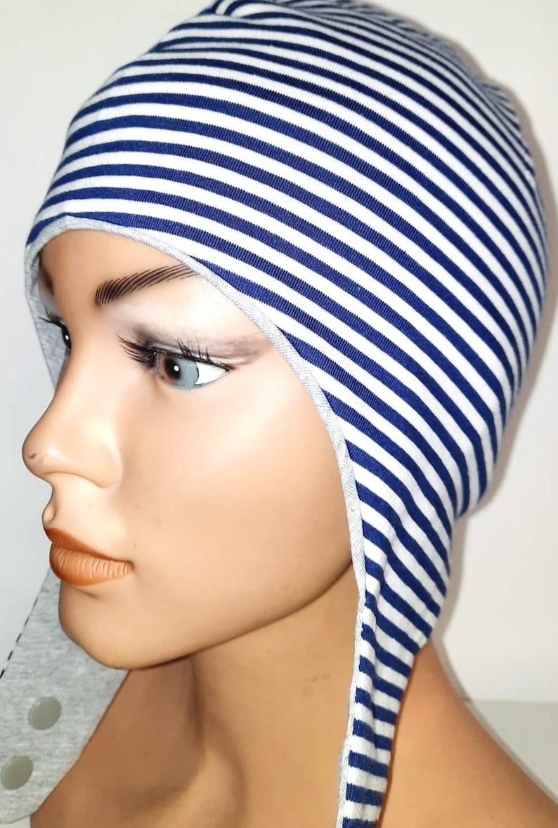 May include: A fitted cap featuring a pattern of thin horizontal navy blue and white stripes. The headwear has extended side flaps that frame the face, finished with a light grey trim along the edges and two visible circular fasteners on the side.