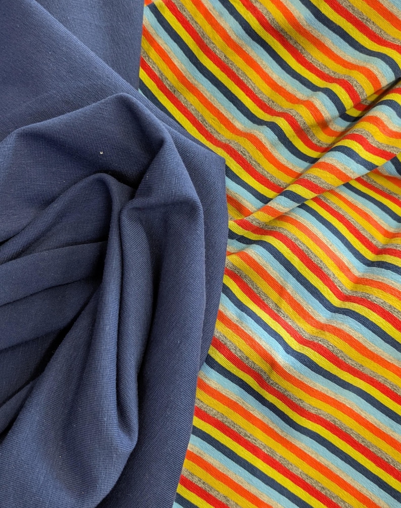 May include: A solid dark navy blue fabric folded next to a multi-coloured striped fabric. The striped fabric features repeating horizontal lines in shades of bright orange, yellow, light blue, and dark navy blue against a heather grey background.