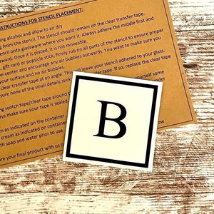May include: Instructions for stencil placement on glassware. The instructions are printed on a brown paper card with a white background. The image also shows a white square with a black border and the letter "B" in black inside the square.