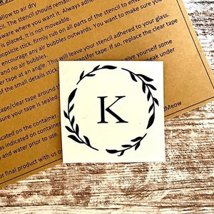 May include: A white sticker with a black wreath design surrounding the letter K. The sticker is on a brown paper background with instructions for using the sticker.