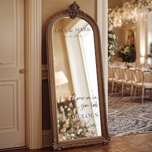 Puede incluir: Un espejo grande y ornamentado con marco dorado con los nombres "LISA & MARK" y la fecha "10.10.2025" en la parte superior. El espejo refleja una habitaci&oacute;n con arreglos florales y el texto "Come on in you look FABULOUS".