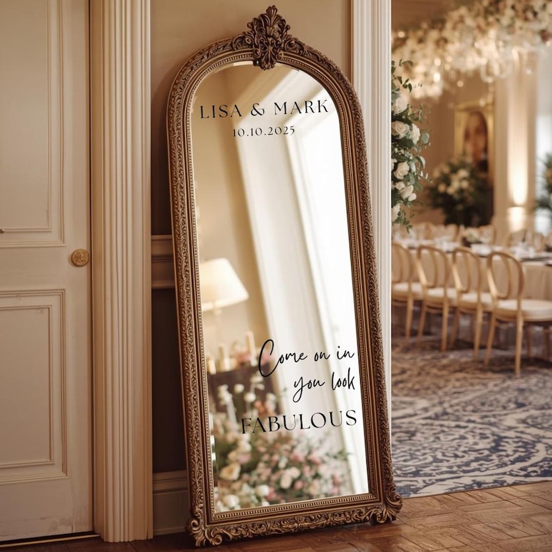 Puede incluir: Un espejo grande y ornamentado con marco dorado con los nombres "LISA & MARK" y la fecha "10.10.2025" en la parte superior. El espejo refleja una habitaci&oacute;n con arreglos florales y el texto "Come on in you look FABULOUS".