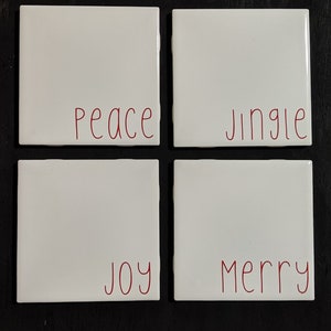 May include: Four white ceramic coasters with red lettering. The words "Peace", "Jingle", "Joy", and "Merry" are written on the coasters.