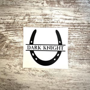 May include: A black horseshoe sticker with the text "DARK KNIGHT" in a white rectangle. The horseshoe has small holes along its curve. The sticker is on a light-colored, textured wooden surface.