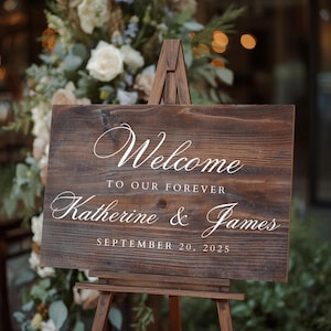 Puede incluir: Un letrero de bienvenida de madera r&uacute;stica sobre un caballete de madera. El letrero dice "Welcome to our forever Katherine & James September 20, 2025" en elegante escritura blanca. El fondo presenta arreglos florales.