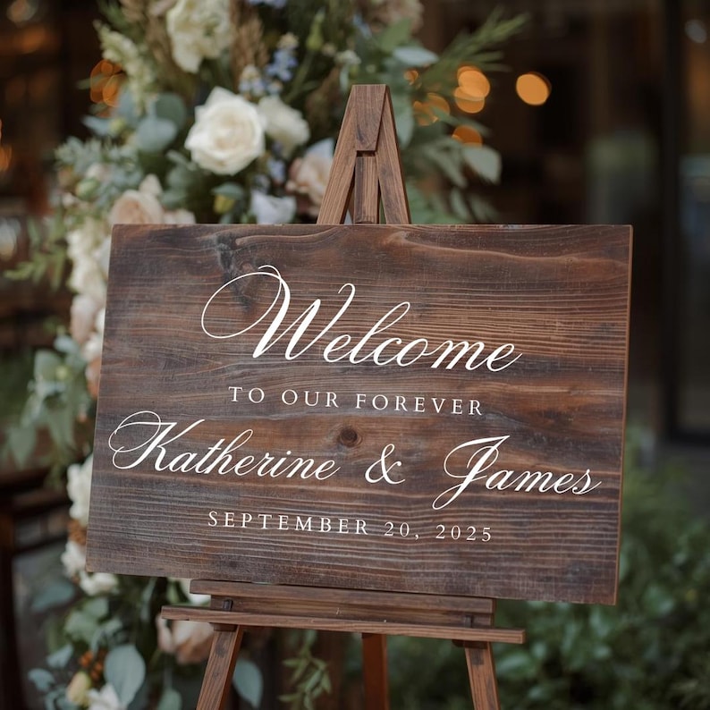 Puede incluir: Un letrero de bienvenida de madera r&uacute;stica sobre un caballete de madera. El letrero dice "Welcome to our forever Katherine & James September 20, 2025" en elegante escritura blanca. El fondo presenta arreglos florales.
