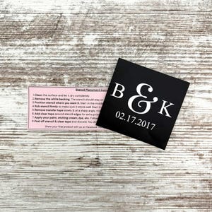 May include: A black square stencil with the letters "B & K" and the date "02.17.2017" in white. A pink instruction sheet is also visible. The stencil is on a light brown wooden surface.