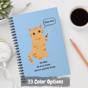 May include: A blue spiral bound notebook with a cartoon orange cat holding a syringe. The text on the notebook says "Come here. Vet Med: We draw blood... and our patients do too."