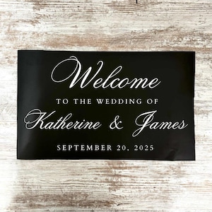 May include: A black sign with white script that reads "Welcome" at the top. Below, it says "To the wedding of Katherine & James" and the date "September 20, 2025". The sign is rectangular.