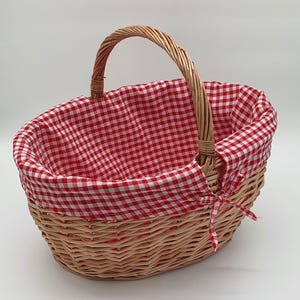 Könnte beinhalten: Ovaler Picknickkorb aus Weide mit rot-weiß kariertem Stofffutter und passender Schleife. Der Korb hat einen geflochtenen Griff und ist ideal für Mahlzeiten im Freien.
