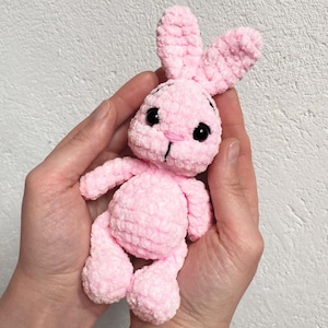 May include: A pink crocheted bunny rabbit toy with black eyes. The bunny is being held in a person's hands.