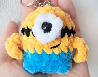 Peut inclure: Un porte-clés en crochet minion jaune et bleu avec un seul œil blanc et des cils noirs. Le porte-clés est attaché à un anneau en métal argenté.