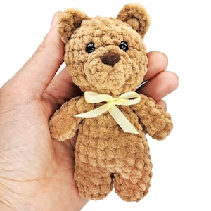 Könnte beinhalten: Ein kleiner brauner gehäkelter Teddybär mit schwarzen Augen und einem gelben Band um den Hals.