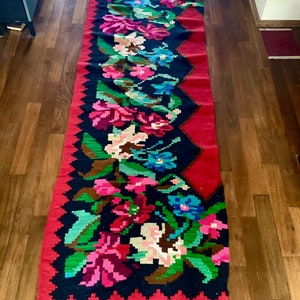 May include: A long, rectangular rug with a red background and a black floral pattern. The rug has a fringe on the bottom edge.
