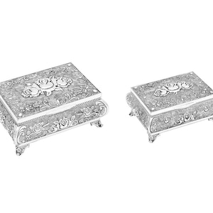 Puede incluir: Dos cajas plateadas ornamentadas con diseños florales de rosas. Las cajas rectangulares tienen tapas con bisagras y se apoyan en pequeños pies. Las tapas presentan un motivo de rosas en relieve y los lados están decorados con patrones florales. Ideales para joyas o recuerdos.