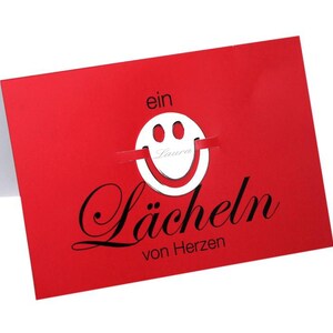 May include: A red card with a white smiley face cutout and the text "ein Lächeln von Herzen" in black.