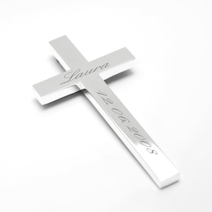 Könnte beinhalten: Ein silbernes Kreuz mit dem Namen "Laura" und dem Datum "12.06.2003" eingraviert.