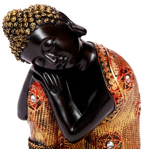 Op de afbeelding: Een zwarte Boeddha-beeld met een serene uitdrukking, die zijn hoofd op zijn hand laat rusten. Het beeld heeft gouden accenten op het haar en de kleding, met rode en gouden patronen en kristallen. Een decoratief stuk voor in huis.