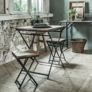 Peut inclure: Une petite table carr&eacute;e et deux chaises pliantes avec des plateaux en bois et des cadres en m&eacute;tal noir. La table est garnie d'assiettes vertes. Une table murale assortie et un seau en bois sont &eacute;galement visibles.