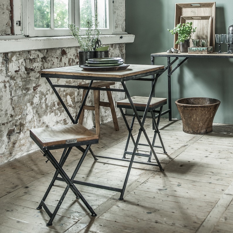 Peut inclure: Une petite table carr&eacute;e et deux chaises pliantes avec des plateaux en bois et des cadres en m&eacute;tal noir. La table est garnie d'assiettes vertes. Une table murale assortie et un seau en bois sont &eacute;galement visibles.