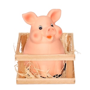 May include: A pink ceramic piggy bank with a smiling face is sitting inside a wooden crate filled with straw.