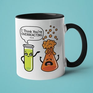 Puede incluir: Taza de cerámica blanca con borde y asa negros. La taza presenta una ilustración de dibujos animados de un tubo de ensayo y un vaso de precipitados con caras. El texto "I Think You're Overreacting" está en un bocadillo de diálogo sobre el tubo de ensayo.