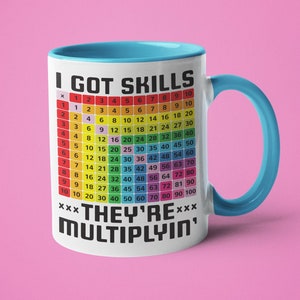 Puede incluir: Taza de cerámica blanca con borde y asa de color azul claro. La taza presenta una tabla de multiplicación de colores arcoíris con el texto "I GOT SKILLS" y "XXX THEY'RE MULTIPLYIN' XXX" en negro.