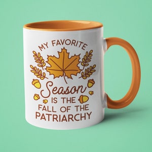 Pode incluir: Caneca de cerâmica branca com borda e alça laranja. A caneca apresenta um gráfico de uma folha de bordo, bolotas e o texto "Minha estação favorita é a queda do patriarcado".