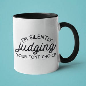 May include: White ceramic mug with a black rim and handle. The mug has black text that reads "I'm silently judging your font choice."