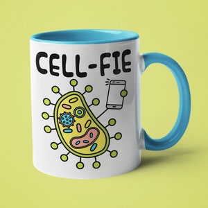 May include: White ceramic mug with a blue rim and handle. The mug features a cartoon illustration of a cell taking a selfie with a smartphone. The text "CELL-FIE" is printed in black on the mug.