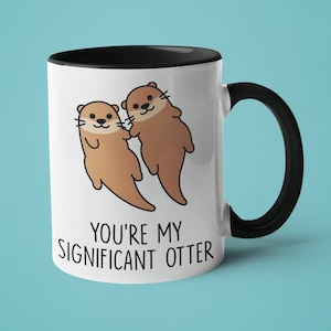 Könnte beinhalten: Eine weiße Keramiktasse mit schwarzem Rand und Henkel. Die Tasse zeigt eine Cartoon-Illustration von zwei Ottern, die sich die Hände halten, und den Text "You're my significant otter".