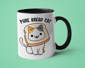 Könnte beinhalten: Eine weiße Keramiktasse mit schwarzem Rand und Henkel. Die Tasse zeigt eine Comic-Katze, die eine Scheibe Brot als Hut trägt. Der Text "PURE BREAD CAT" ist auf die Tasse gedruckt.