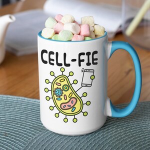May include: White ceramic mug with a blue handle. The mug features a cartoon cell with a phone taking a selfie. The text on the mug reads "CELL-FIE".
