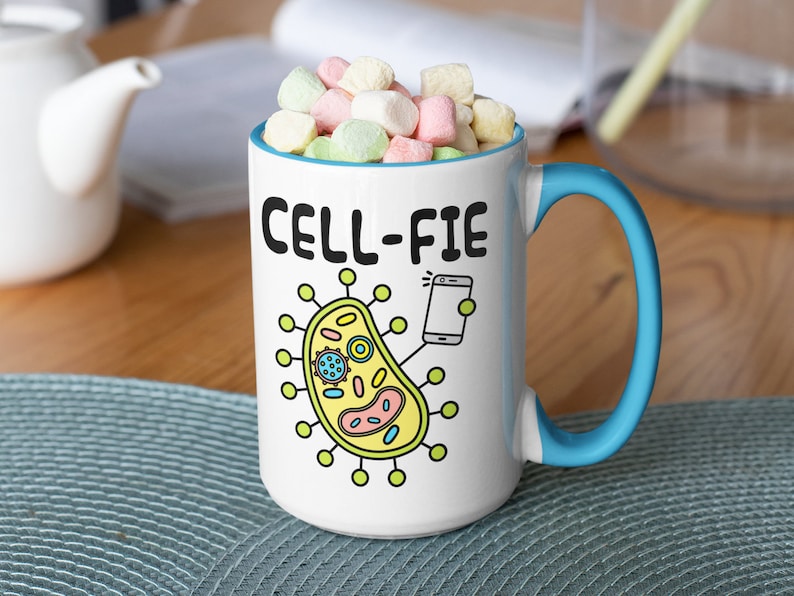 May include: White ceramic mug with a blue handle. The mug features a cartoon cell with a phone taking a selfie. The text on the mug reads "CELL-FIE".