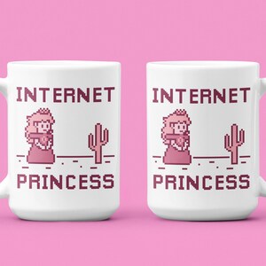K&ouml;nnte beinhalten: Wei&szlig;e Keramiktasse mit dem Text "INTERNET PRINCESS" in einer pixeligen Schriftart, zusammen mit einem pixeligen Bild einer Prinzessin und eines Kaktus. Die Tasse hat einen Henkel und steht vor einem rosa Hintergrund.