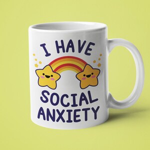 May include: White ceramic mug with a rainbow and two yellow stars with faces. The text "I have social anxiety" is printed on the mug.