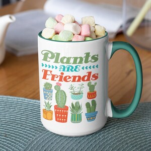 May include: White ceramic mug with a green handle. The mug has a colorful design of potted cacti and the text "Plants are Friends".