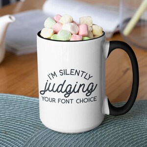Puede incluir: Taza de cer&aacute;mica blanca con asa y borde negros. La taza tiene un gr&aacute;fico negro que dice "Estoy juzgando silenciosamente tu elecci&oacute;n de fuente".
