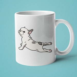 May include: White ceramic coffee mug with a cartoon illustration of a French bulldog doing yoga. The bulldog is grey and white and is in a downward-facing dog pose.