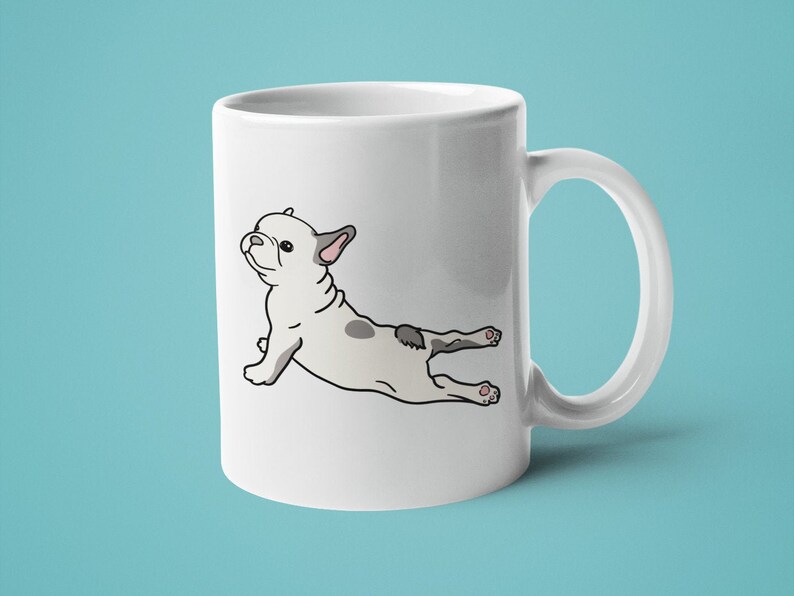 May include: White ceramic coffee mug with a cartoon illustration of a French bulldog doing yoga. The bulldog is grey and white and is in a downward-facing dog pose.