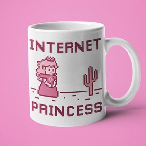 K&ouml;nnte beinhalten: Wei&szlig;e Keramik-Tasse mit dem Text "INTERNET PRINCESS" in pixeligen, kastanienbraunen Buchstaben. Die Tasse zeigt eine pixelige Illustration einer Prinzessin und eines Kaktus. Die Tasse hat einen wei&szlig;en Henkel und steht vor einem rosa Hintergrund.