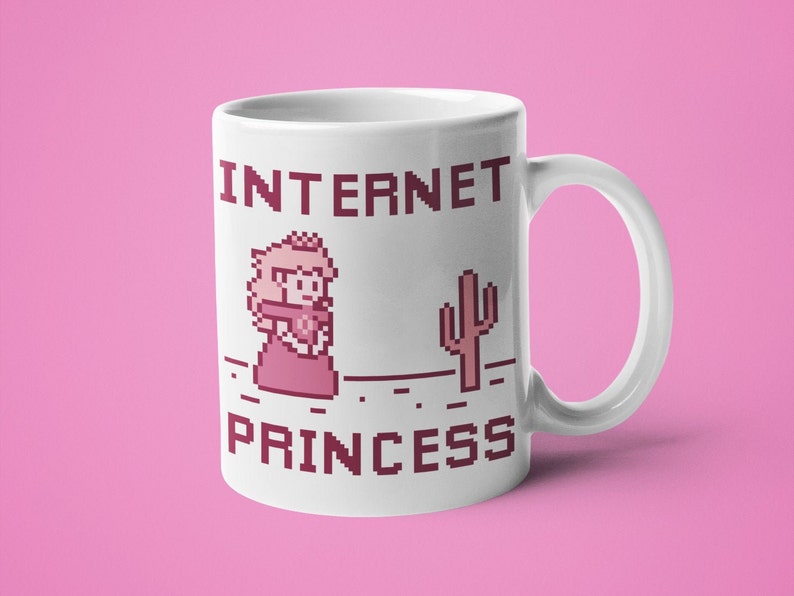 K&ouml;nnte beinhalten: Wei&szlig;e Keramik-Tasse mit dem Text "INTERNET PRINCESS" in pixeligen, kastanienbraunen Buchstaben. Die Tasse zeigt eine pixelige Illustration einer Prinzessin und eines Kaktus. Die Tasse hat einen wei&szlig;en Henkel und steht vor einem rosa Hintergrund.